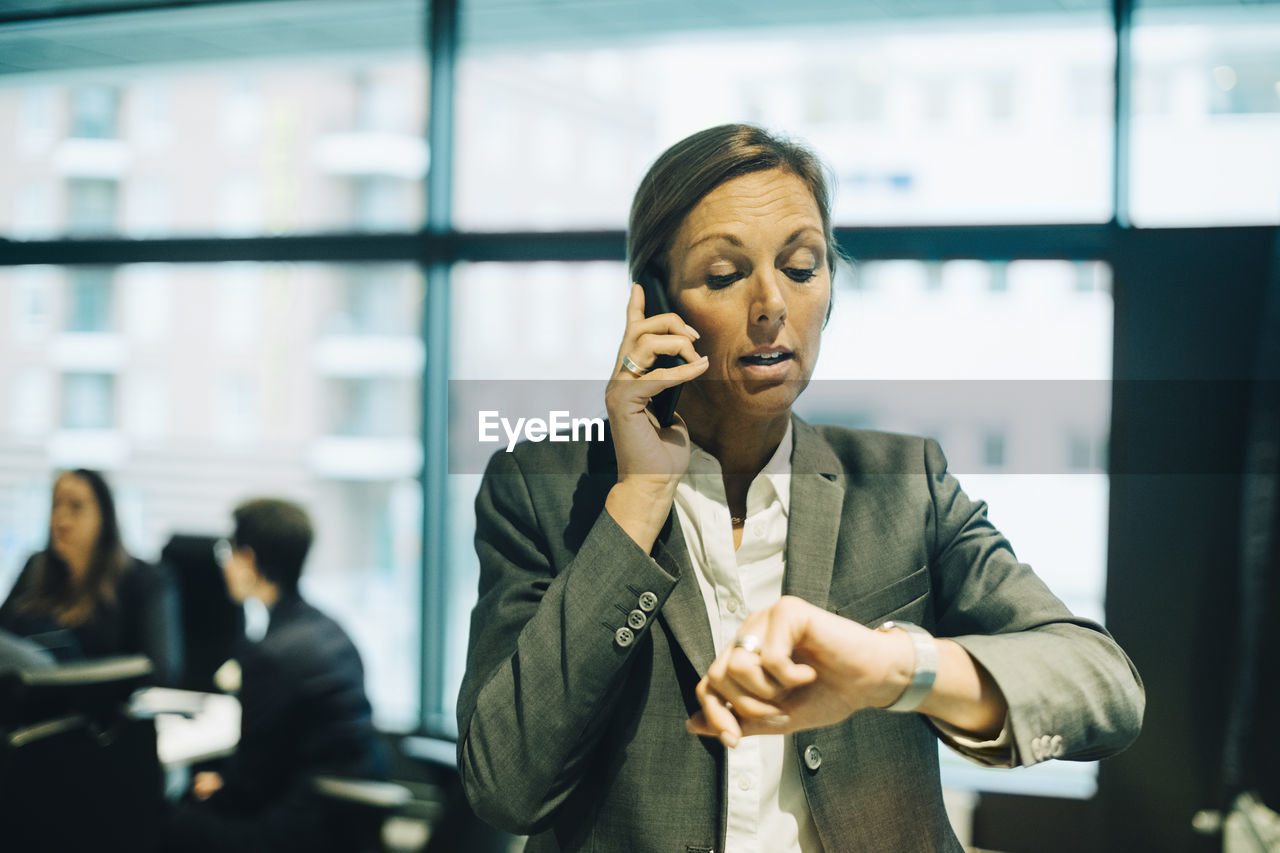 Busy businesswoman talking on mobile phone while checking time on watch at office