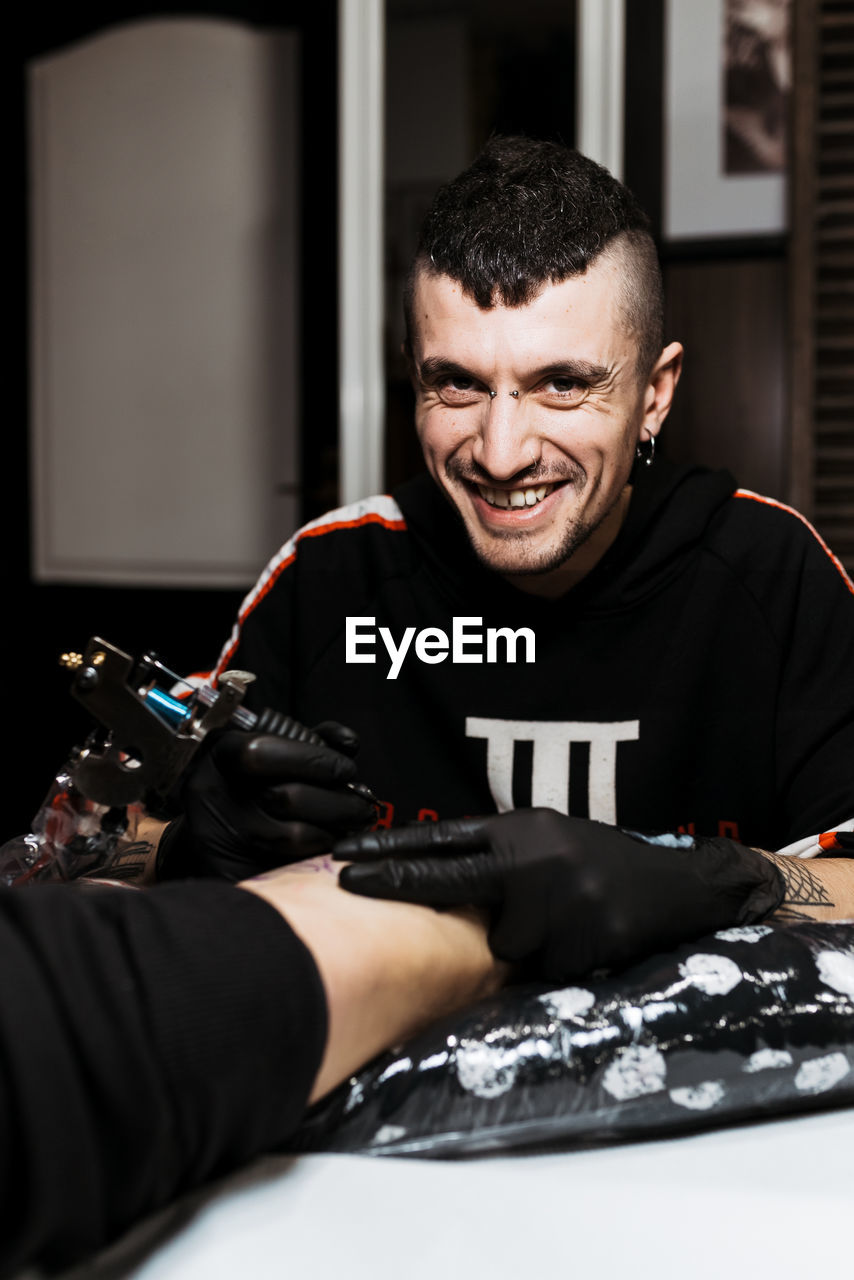 Stylish happy man with piercing looking at camera while using tattoo machine to make tattoo on leg of crop anonymous customer during work in salon