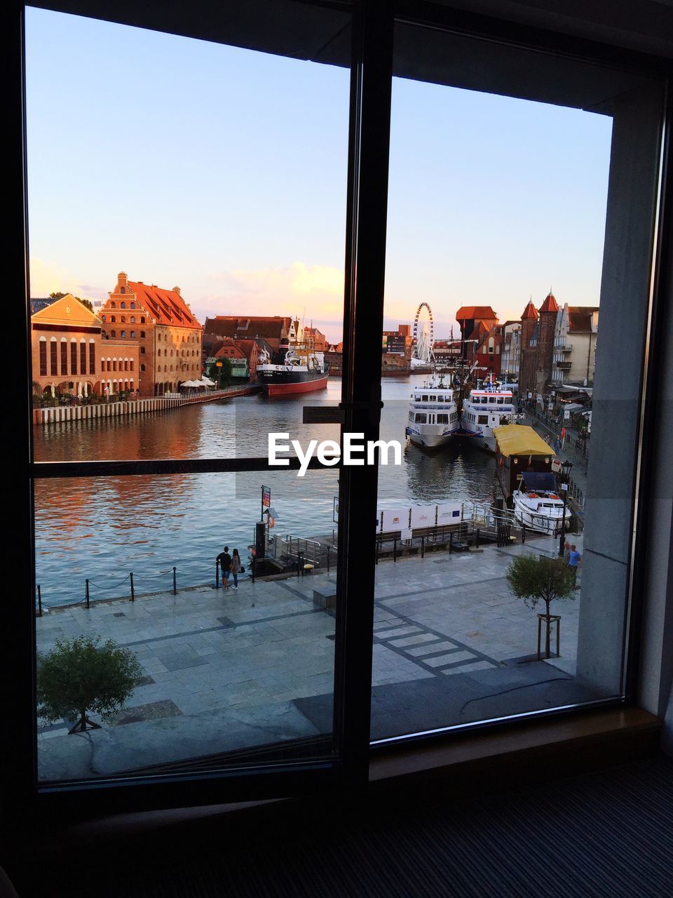 Canal amidst buildings in front of ferris wheel seen through glass window against sky