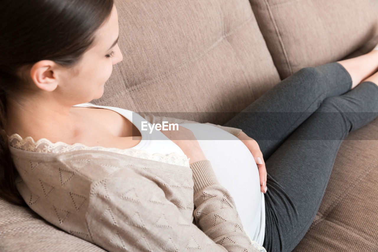 Photo of cute caucasian future mom lying in light living room on sofa and holding hands on tummy.