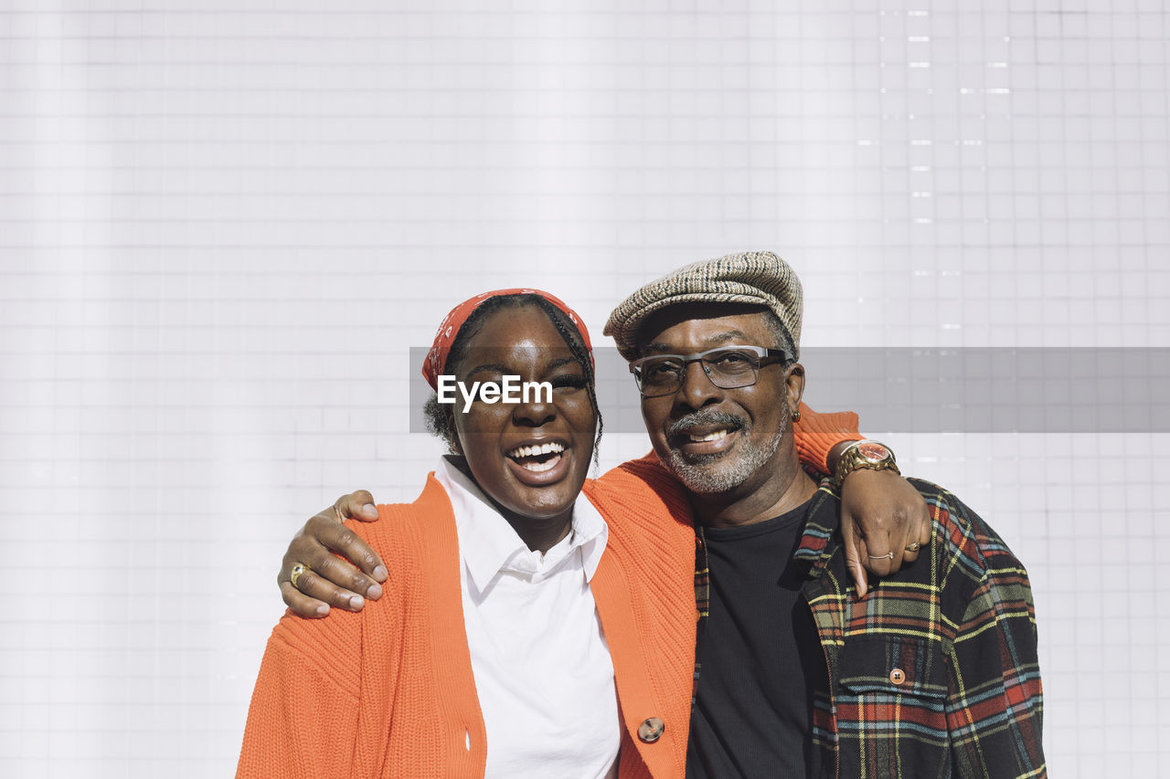 Portrait of cheerful father and daughter with arms around against wall during sunny day