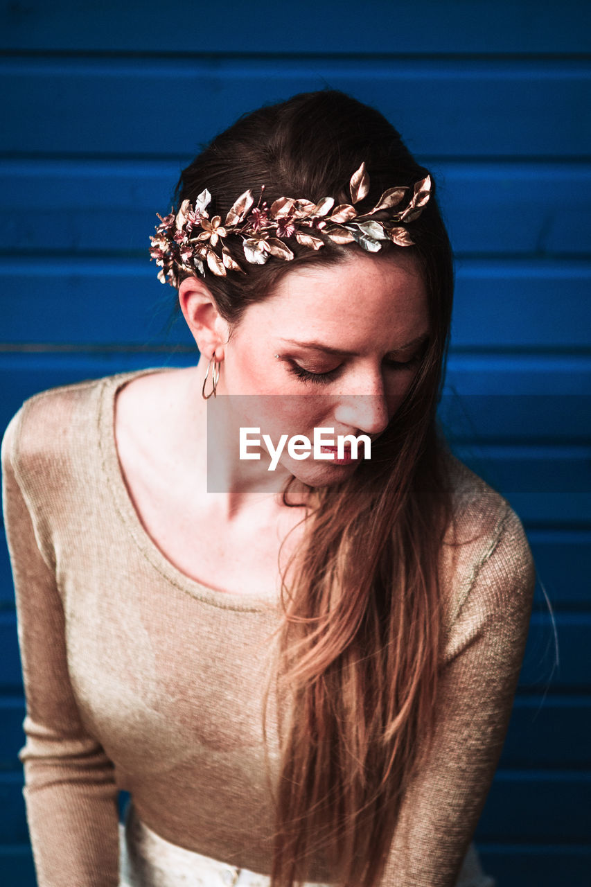 Tender calm young female in trendy wear and copper headband standing against blue plank wall and looking down