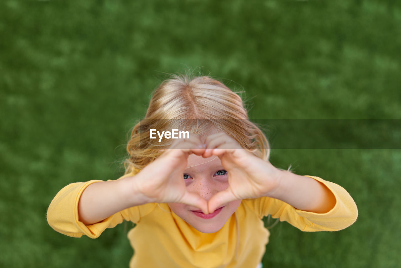 Pretty blonde child girl showing sign of love heart from fingers hands. top view