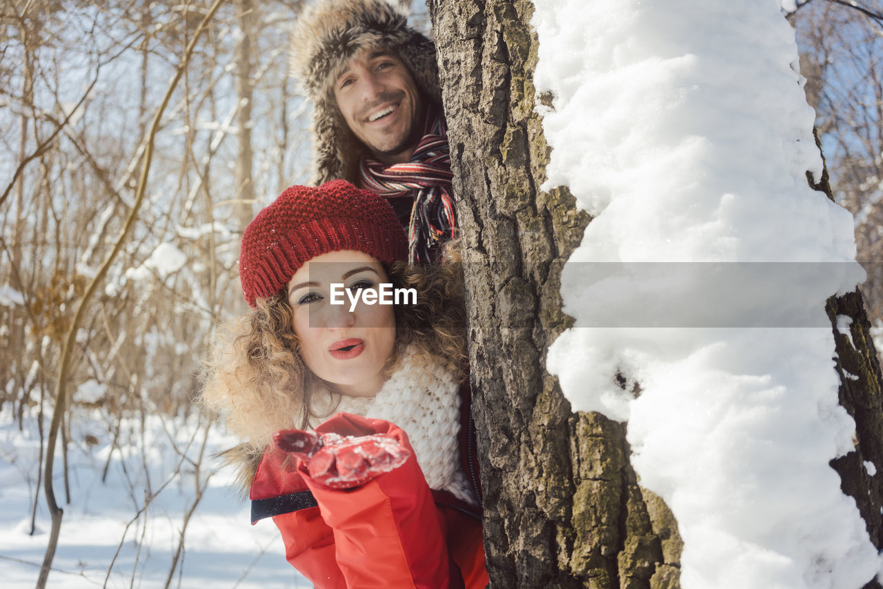 Portrait of smiling couple peeking through tree trunk in forest during winter