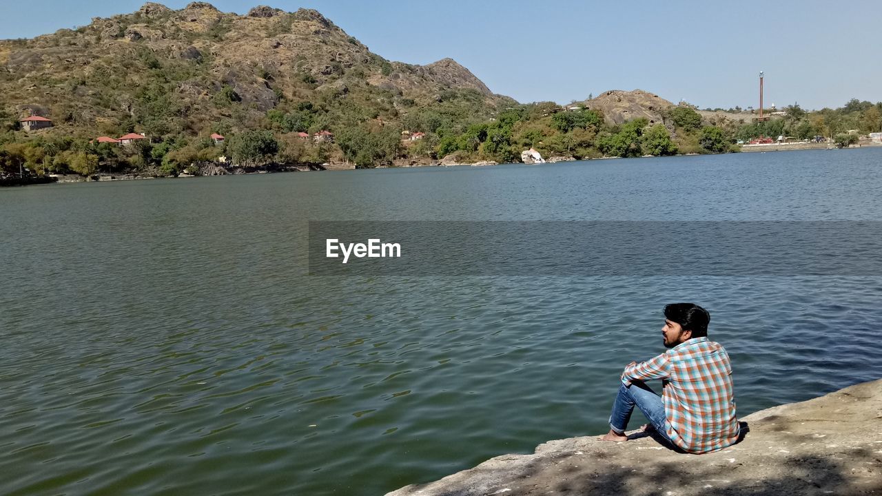 Rear view of young man sitting on rock by lake against sky