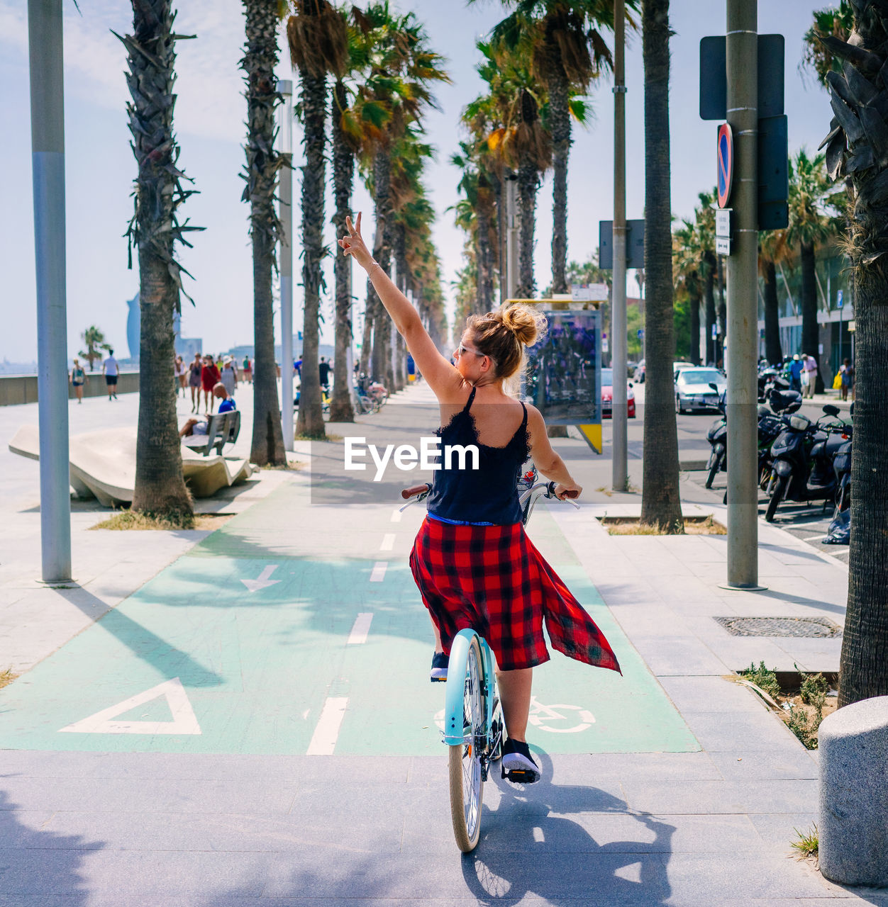 Back view of cool female bicyclist riding bike while demonstrating victory gesture with raised arm on city walkway