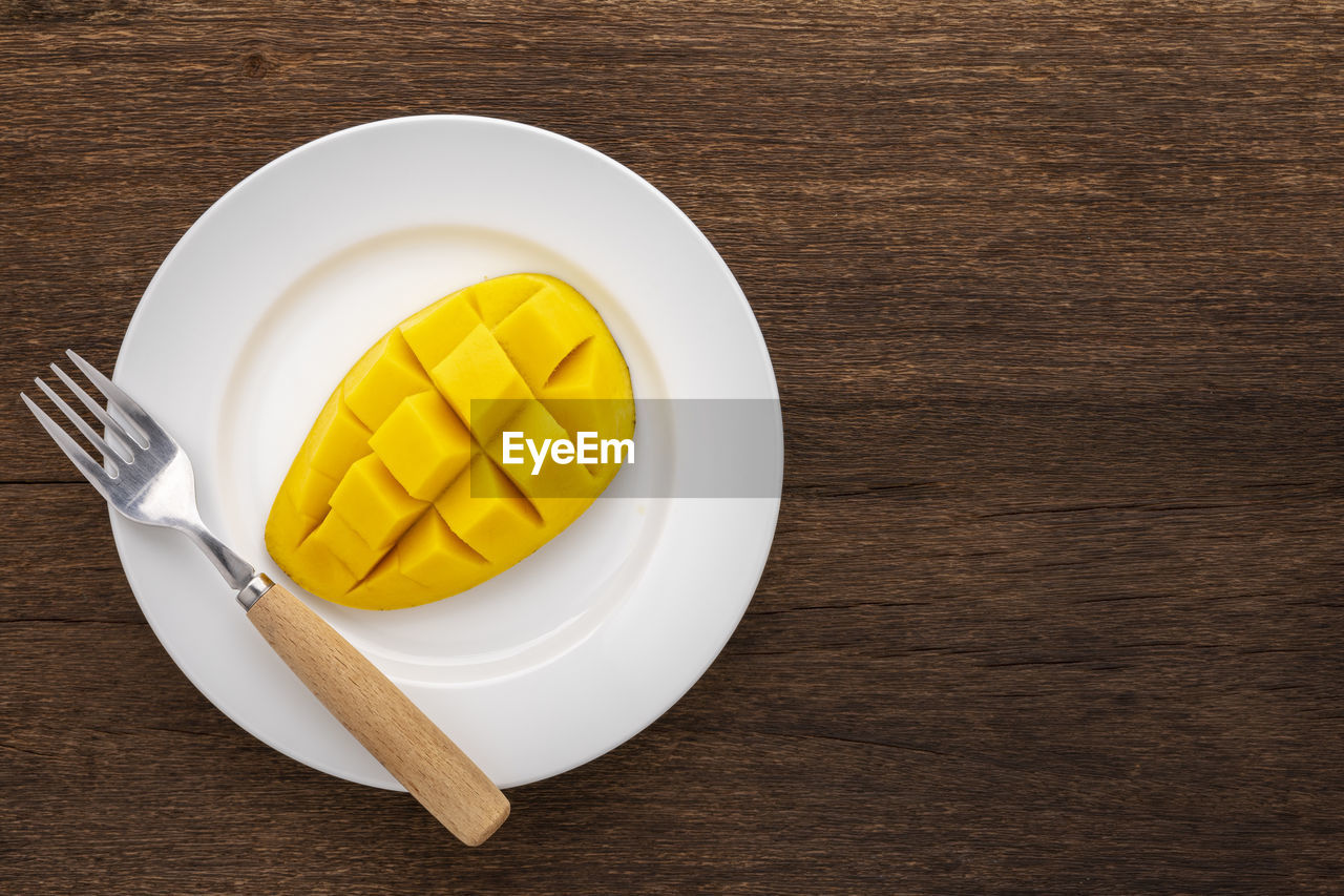 Tasty fresh yellow ripe mango with fork in simply white ceramic plate on rustic wood, top view 