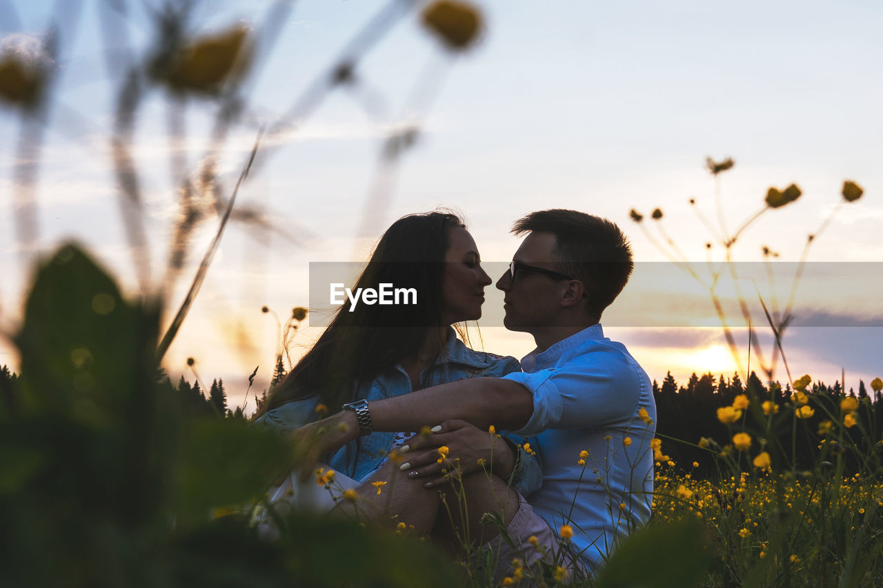 Couple sitting amidst flowering plants against sky during sunset