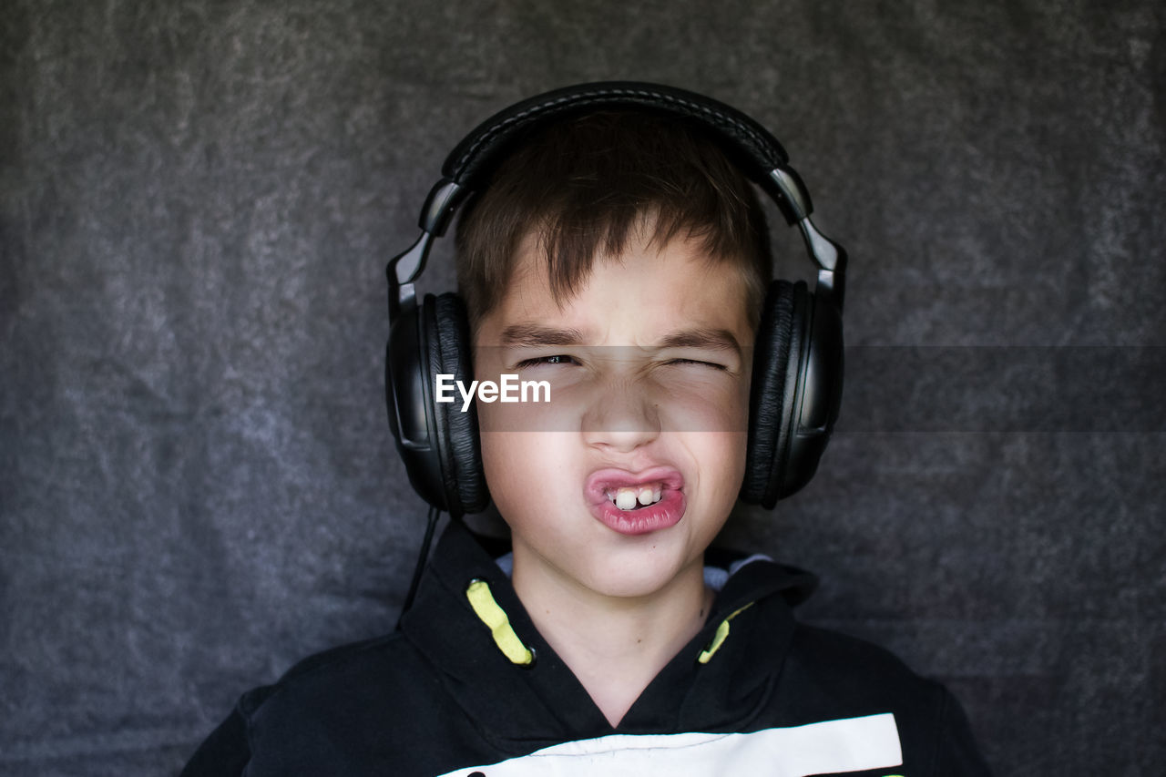 Boy making face while listening music in headphones against wall