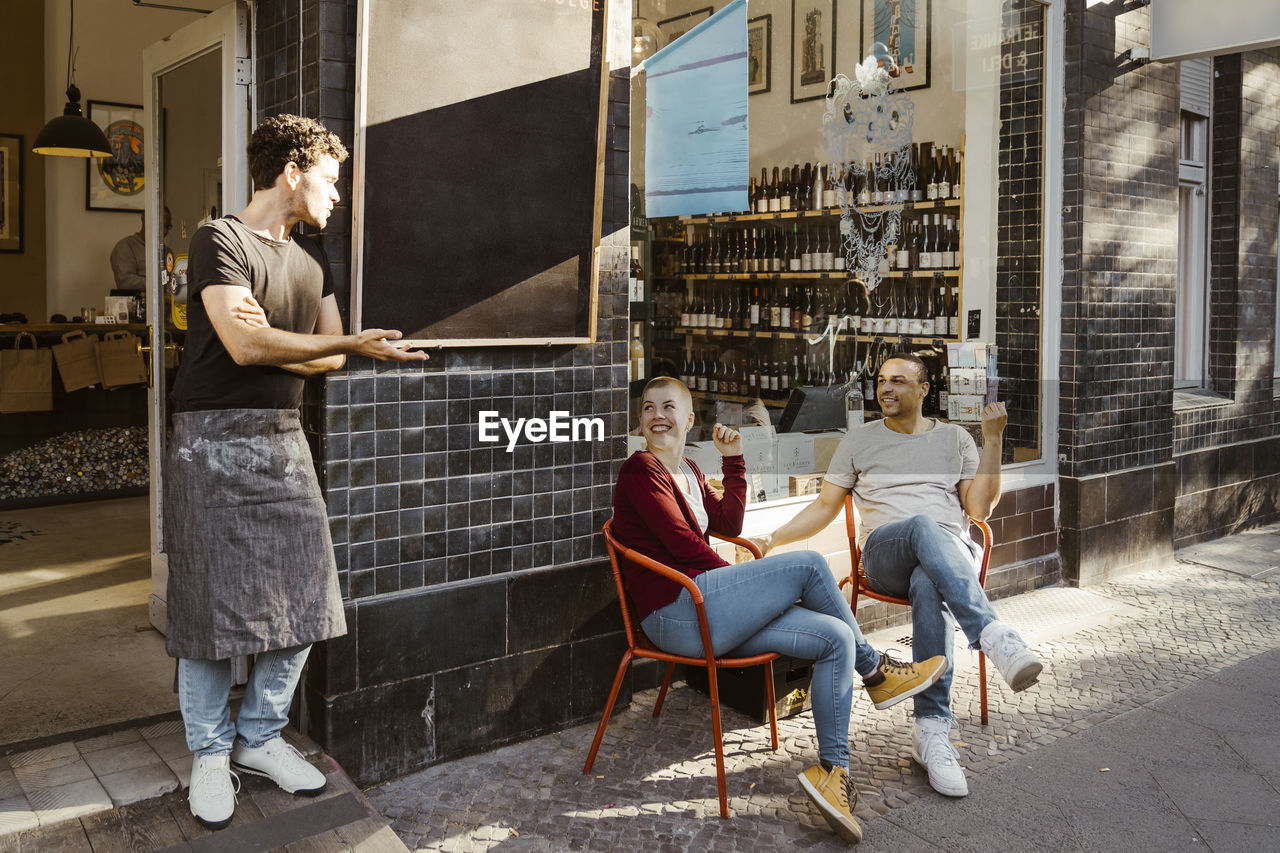 Male owner talking to customers sitting on chair at sidewalk outside store