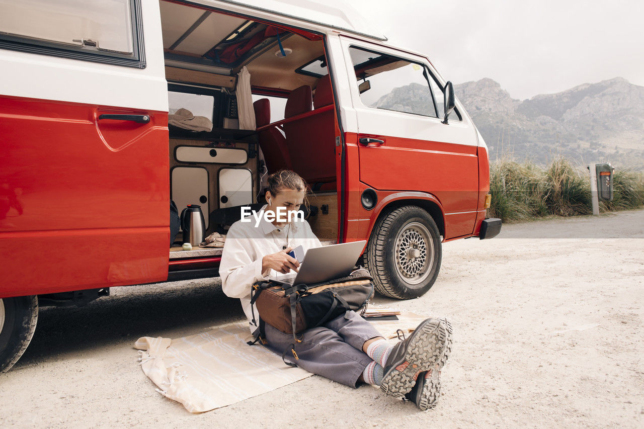 Young man using laptop while sitting on blanket near van during vacation