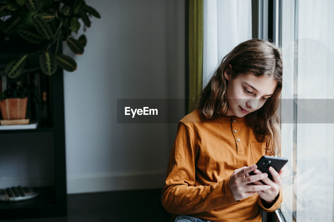 Girl using mobile phone while leaning on window at home