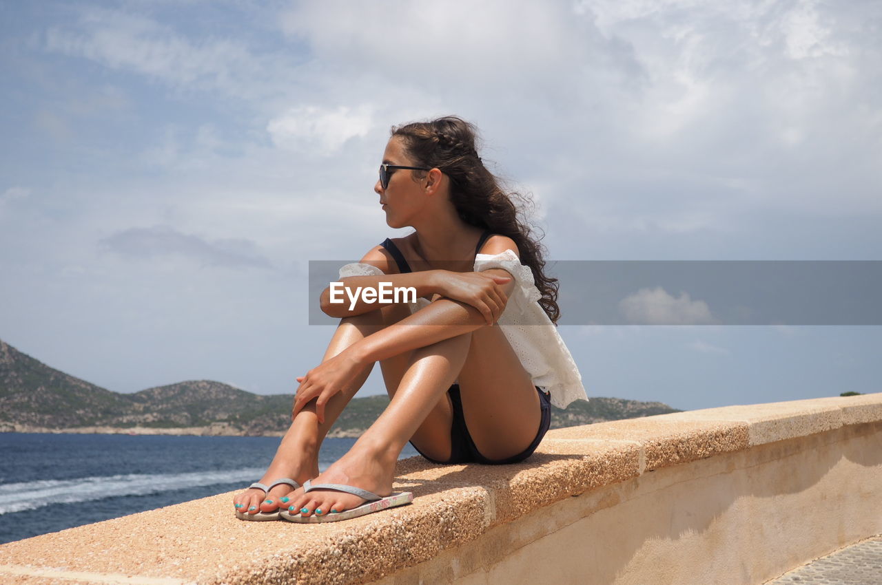 Full length of young woman relaxing on retaining wall by sea