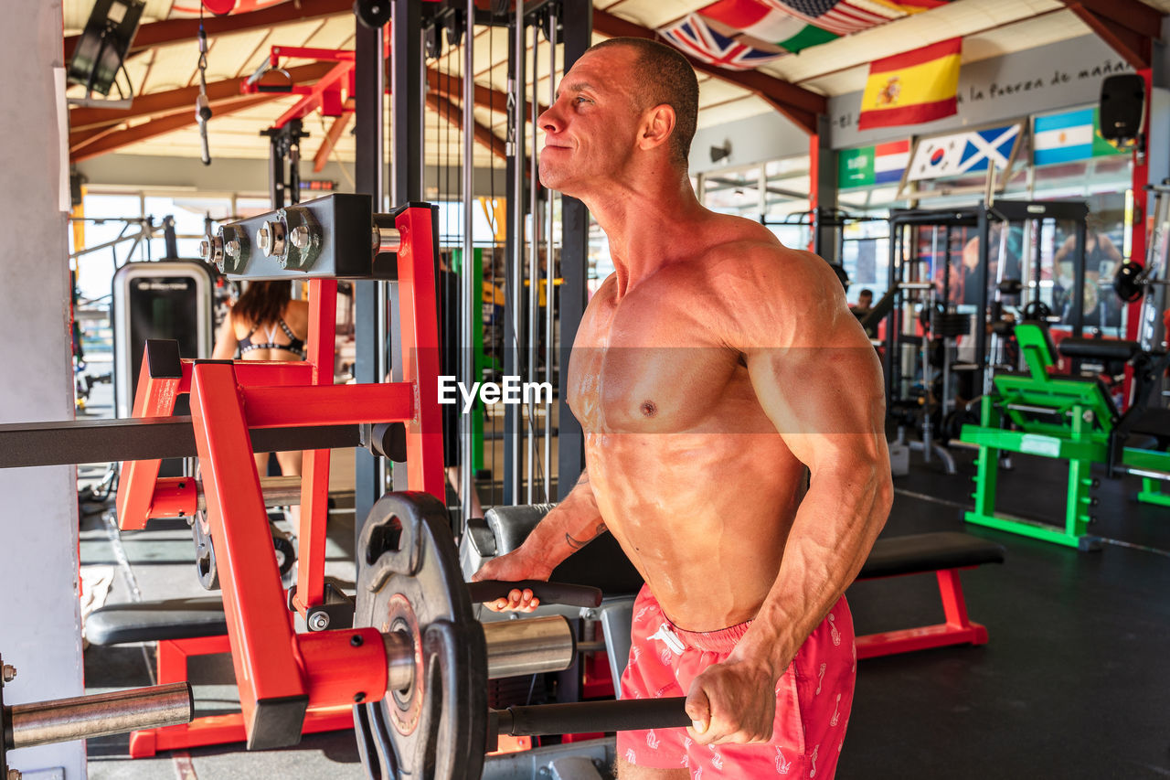 Side view of powerful male athlete with sweaty torso lifting heavy barbell plate during workout in sports club