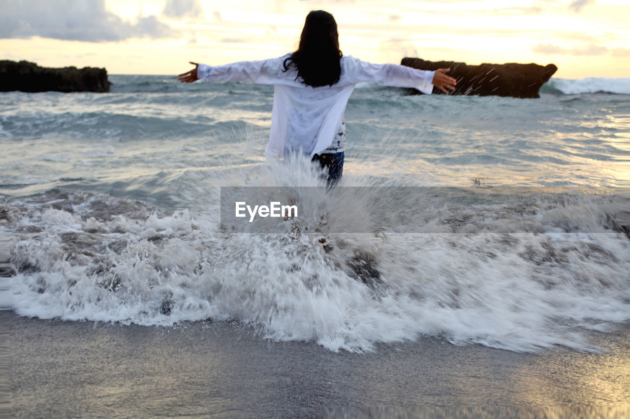 Rear view of teenage girl with arms outstretched standing at beach against sky during sunset