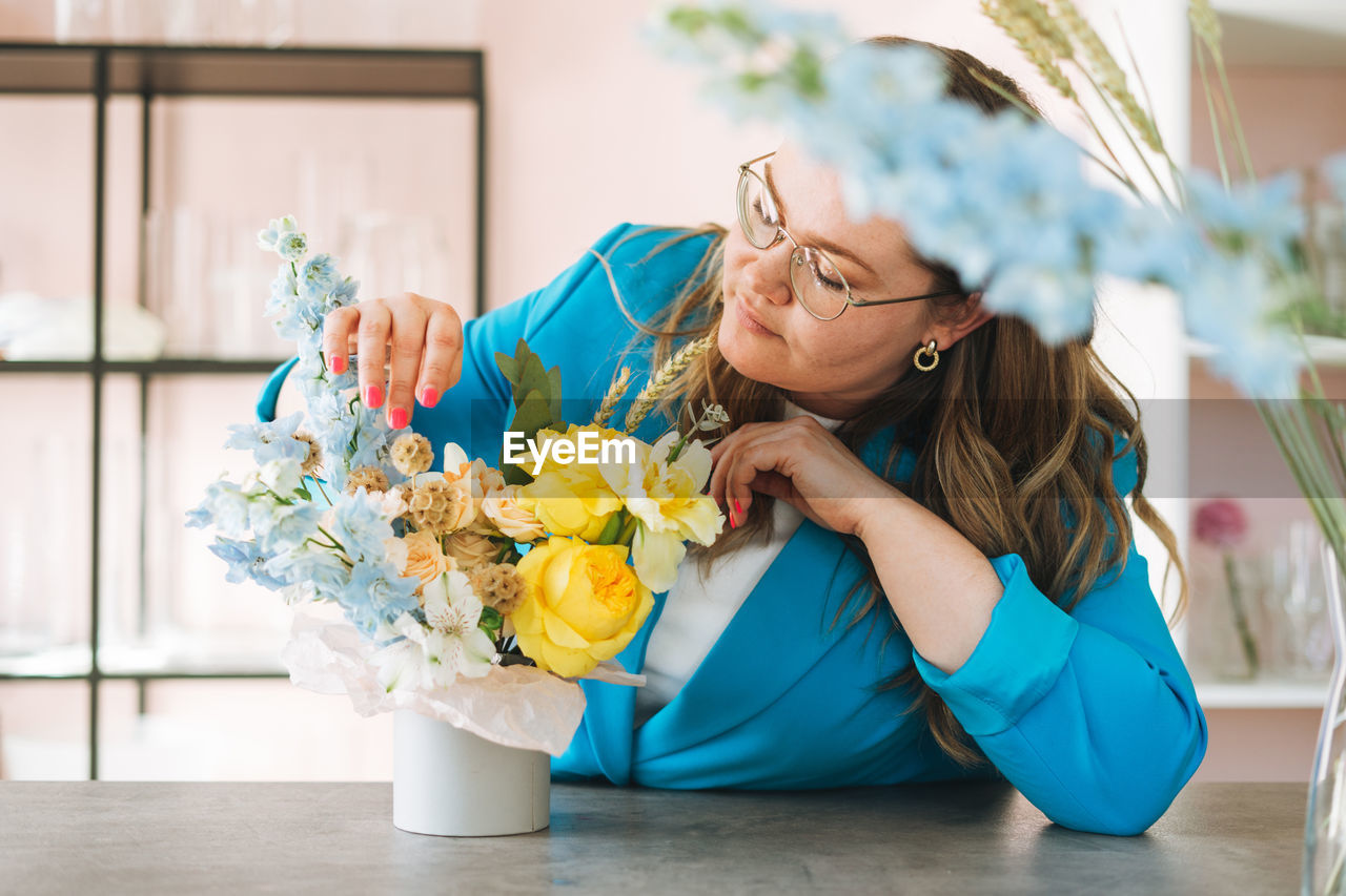 Young woman florist in blue suit and eye glasses with bouquet of flowers in box in flower shop