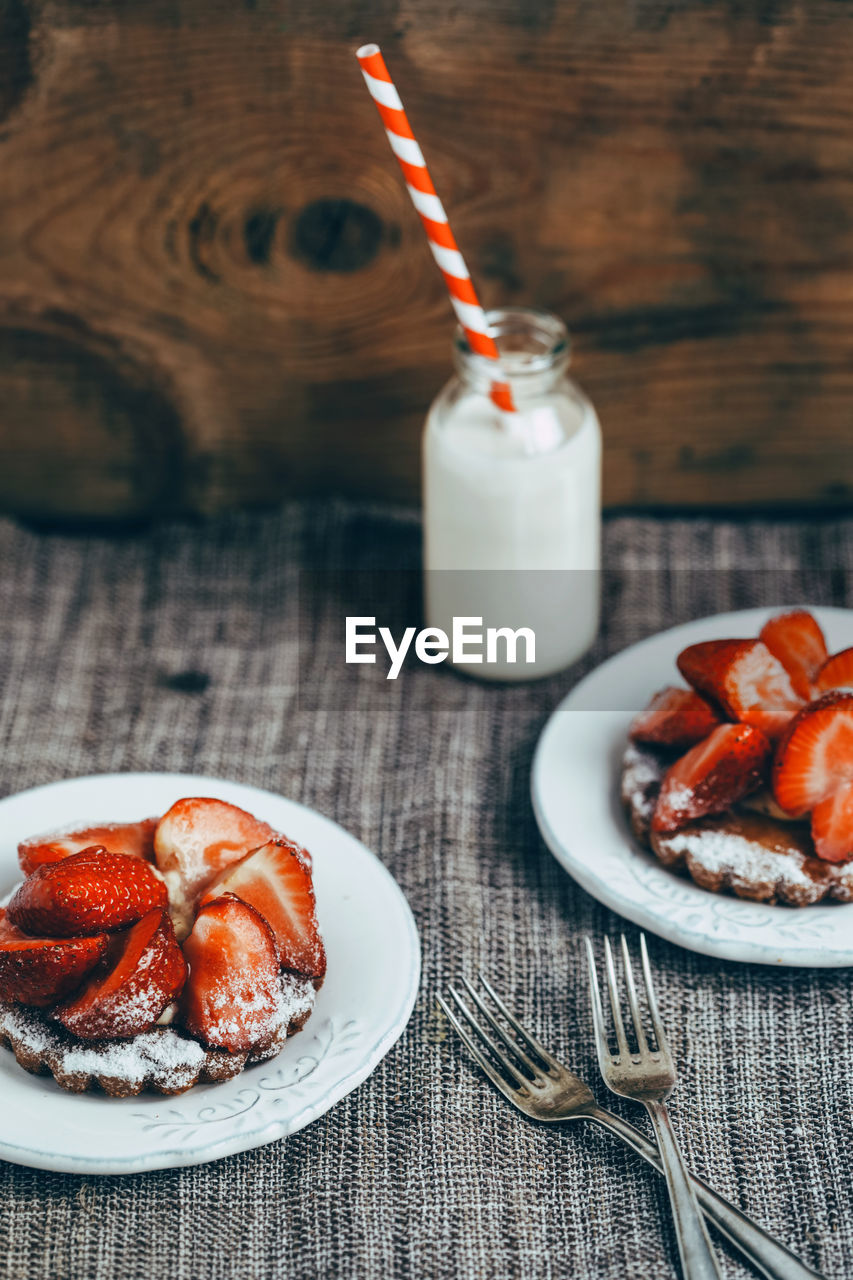 From above of delicious homemade waffles with ripe strawberry and sugar powder served on white plates on table with milk drink in glass bottle