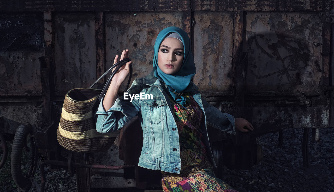 Young woman holding bag while sitting on abandoned machinery