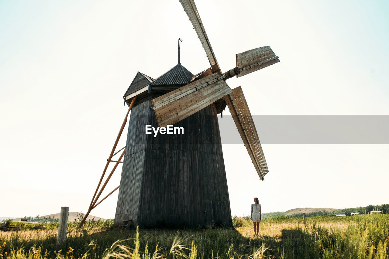 Woman standing by traditional windmill on field against clear sky