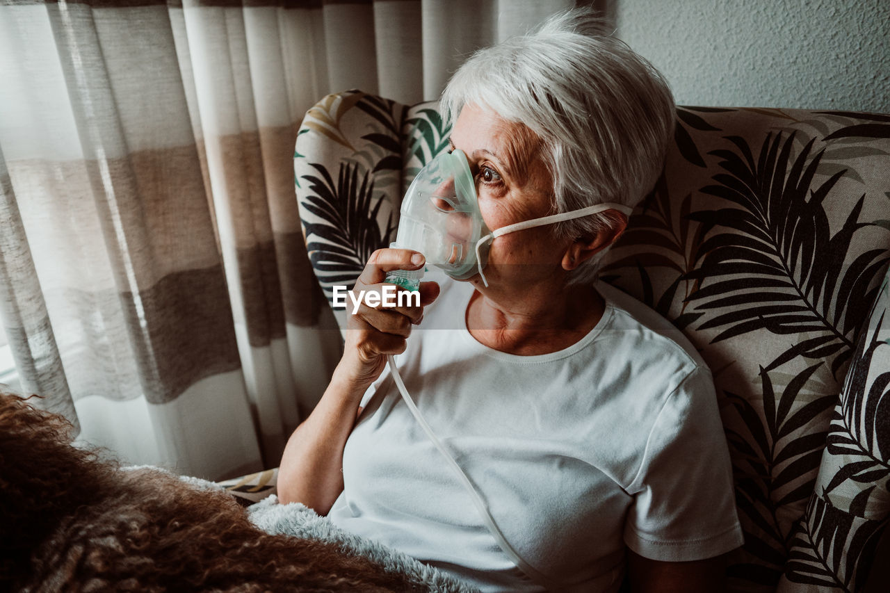 Senior woman wearing oxygen mask sitting with dog on chair at home