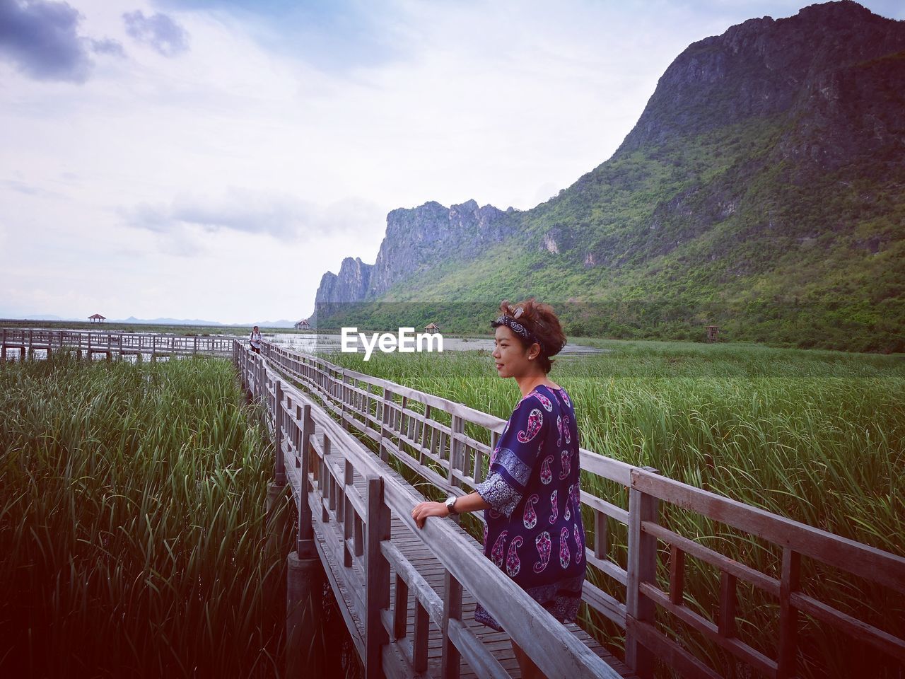 Thoughtful woman standing on wooden footbridge over grassy field against sky