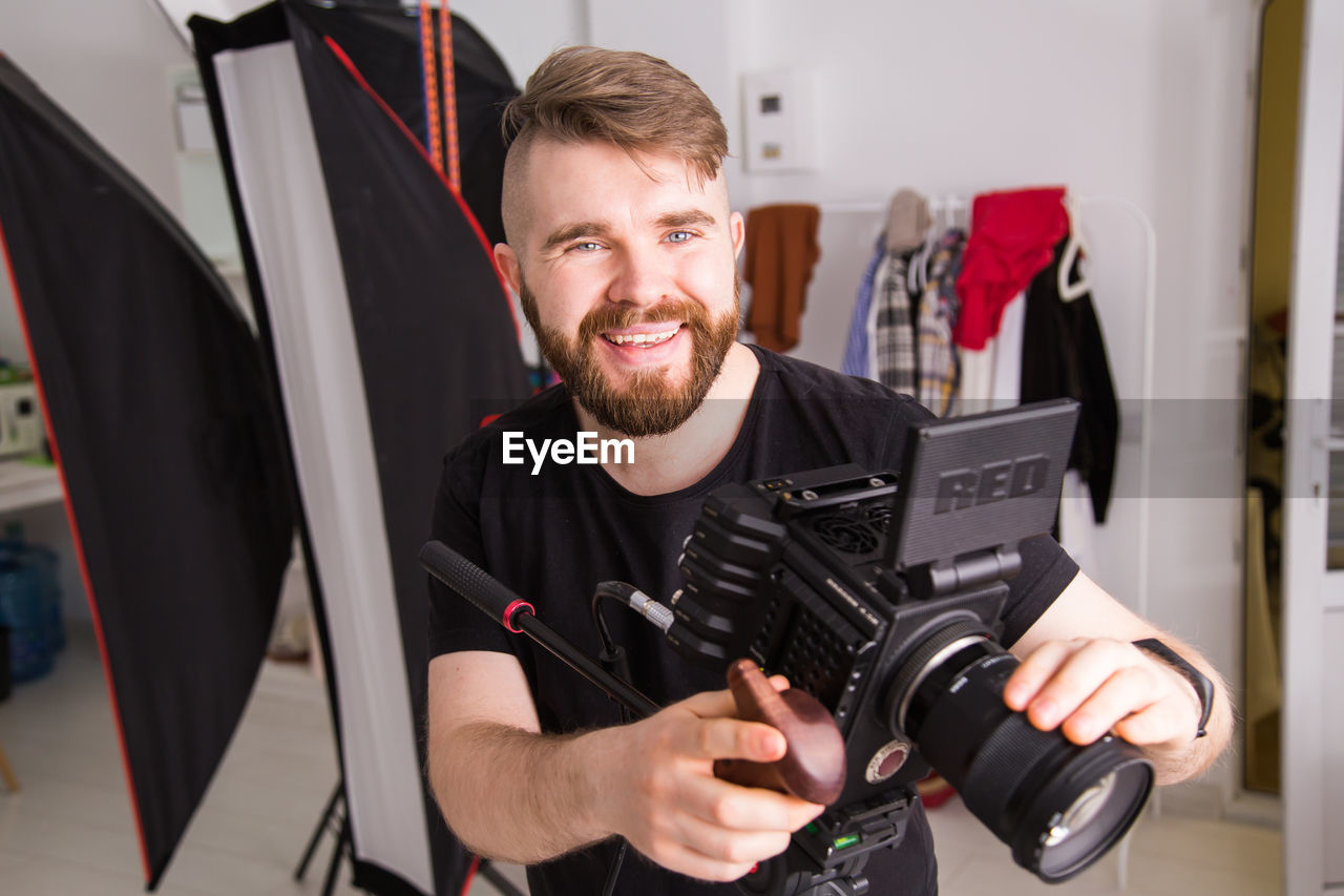 one person, adult, occupation, men, smiling, beard, indoors, technology, facial hair, portrait, working, front view, happiness, camera, expertise, looking at camera, skill, business finance and industry, person, casual clothing, arts culture and entertainment, clothing, young adult, business, small business, standing, studio, room, emotion, holding, waist up, creativity, equipment, tripod, filmmaking