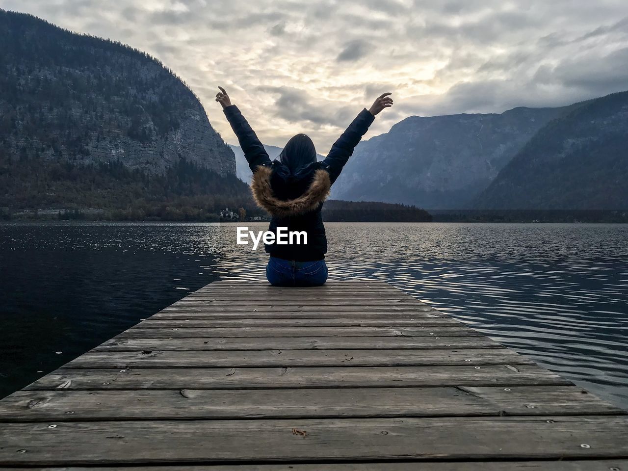 Rear view of woman with arms raised sitting on pier over lake against sky