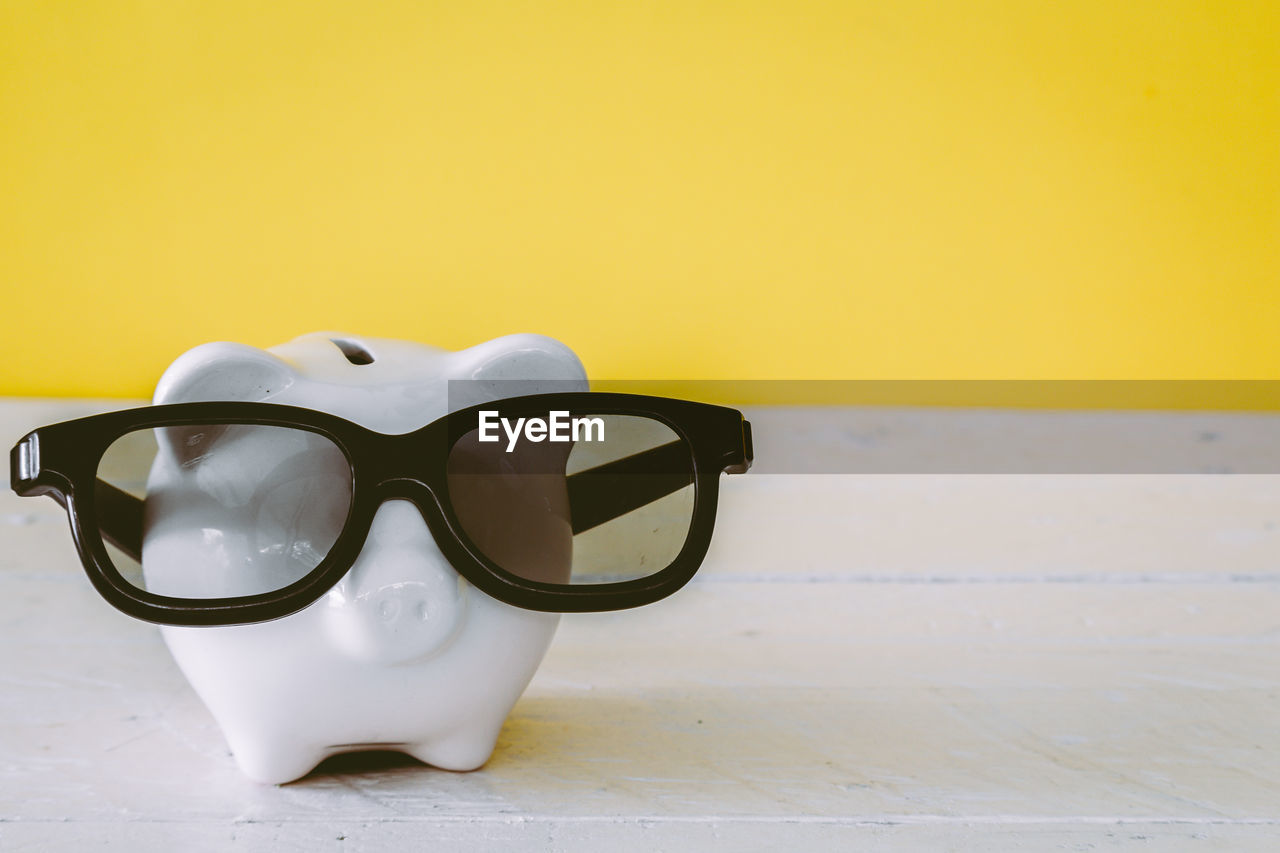 Close-up of sunglasses with piggy bank on table against yellow wall
