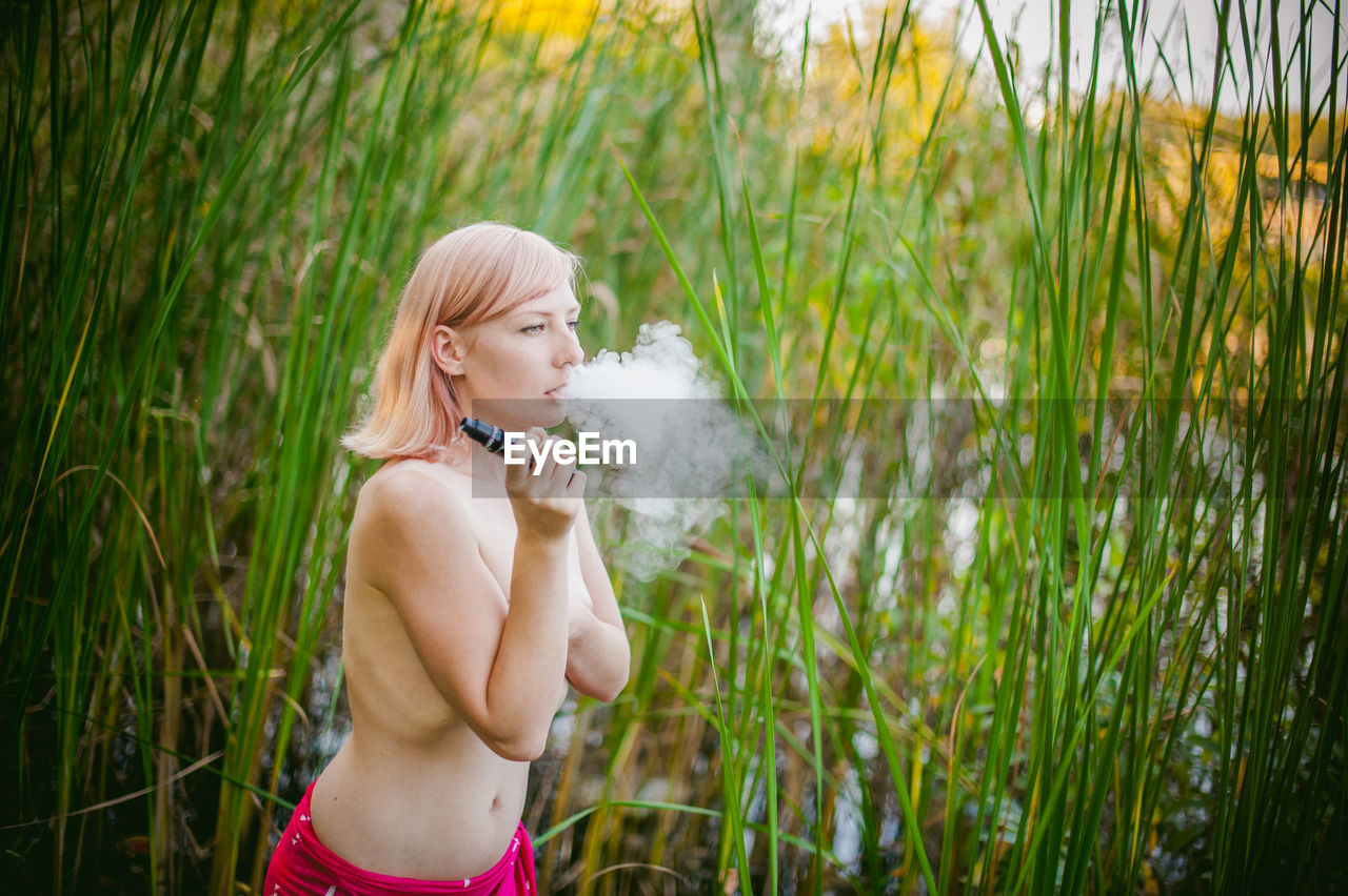 Rear view of shirtless young woman smoking while standing on grassy field