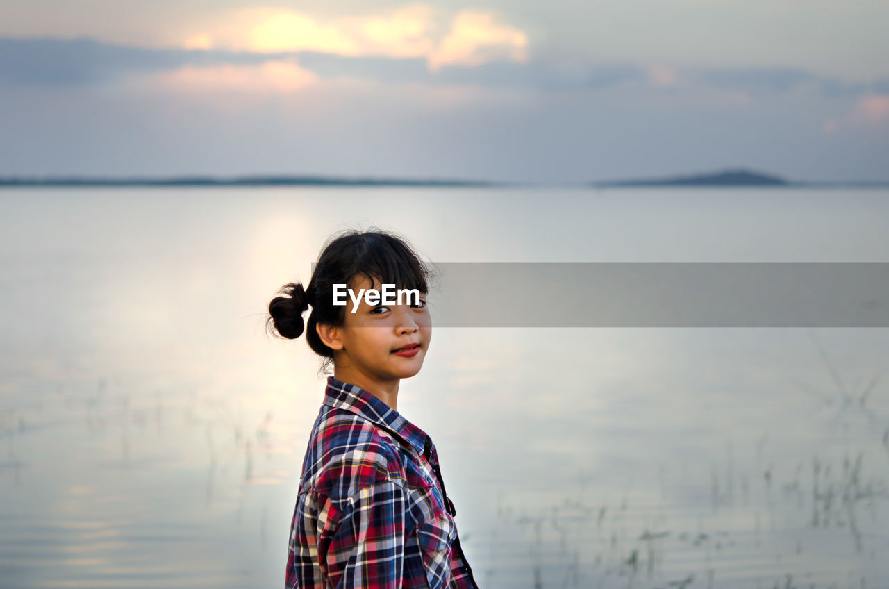 Side view portrait of girl standing against lake during sunset