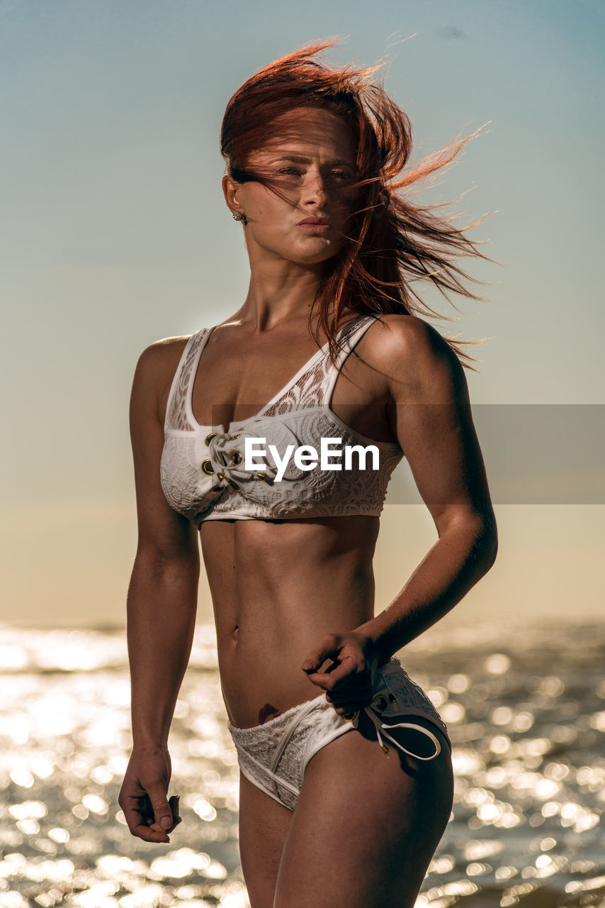 Sensuous female model standing in bikini at beach against sky