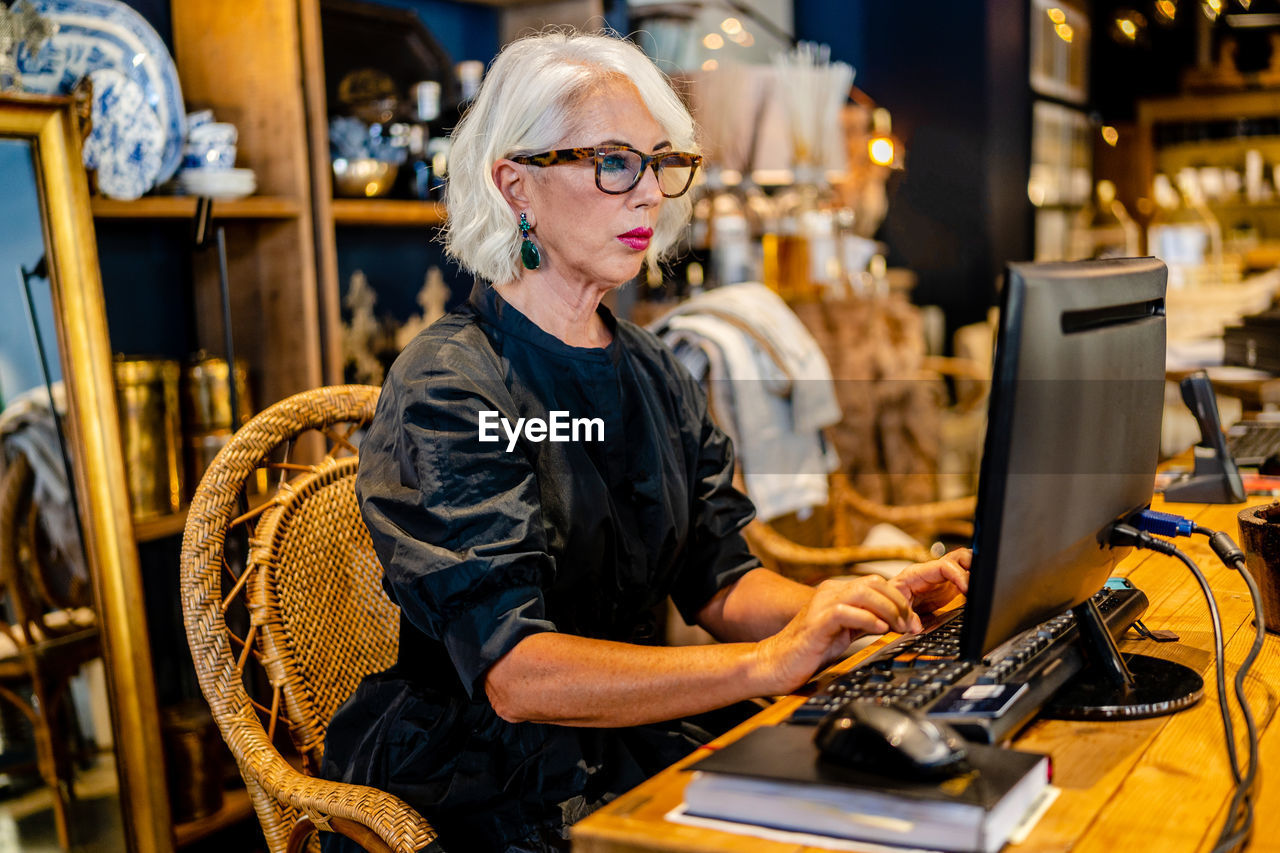 Side view of mature female entrepreneur in elegant dress sitting at table and typing on keyboard while working on computer on project online