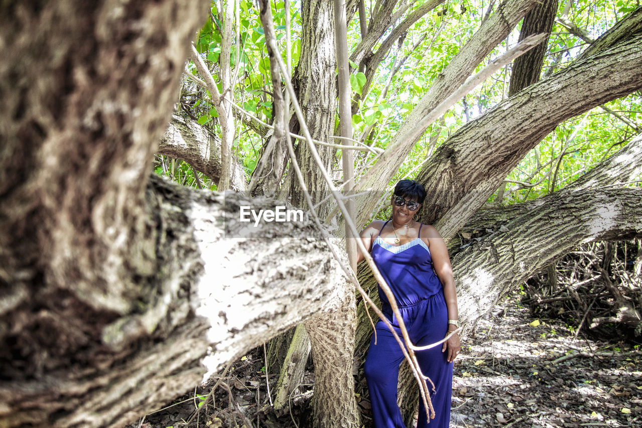 Smiling woman wearing sunglasses standing by tree trunk in forest