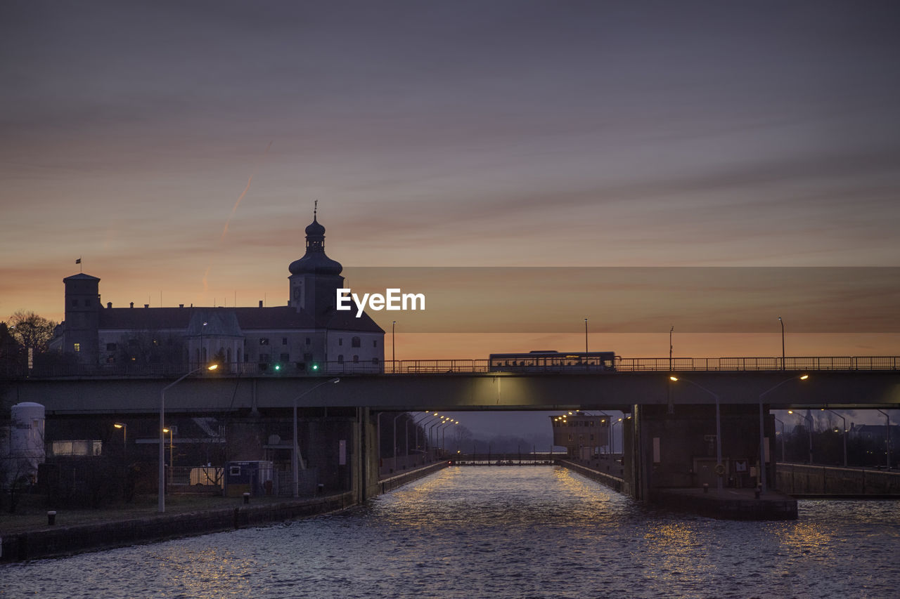 ILLUMINATED BRIDGE OVER RIVER BY BUILDINGS AGAINST SKY AT SUNSET