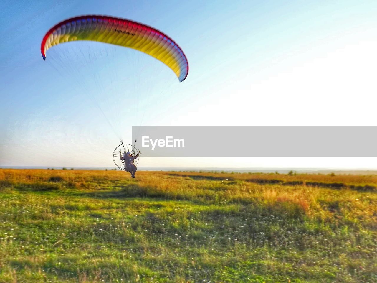 Man flying powered parachute over grassy field against clear sky