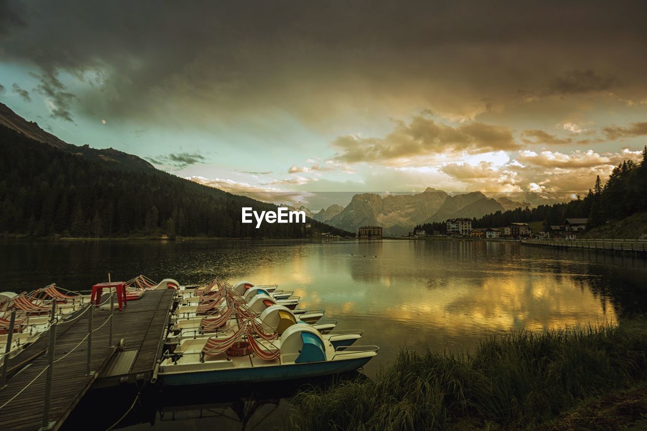 Boats moored at lake misurina by mountains against cloudy sky during sunset