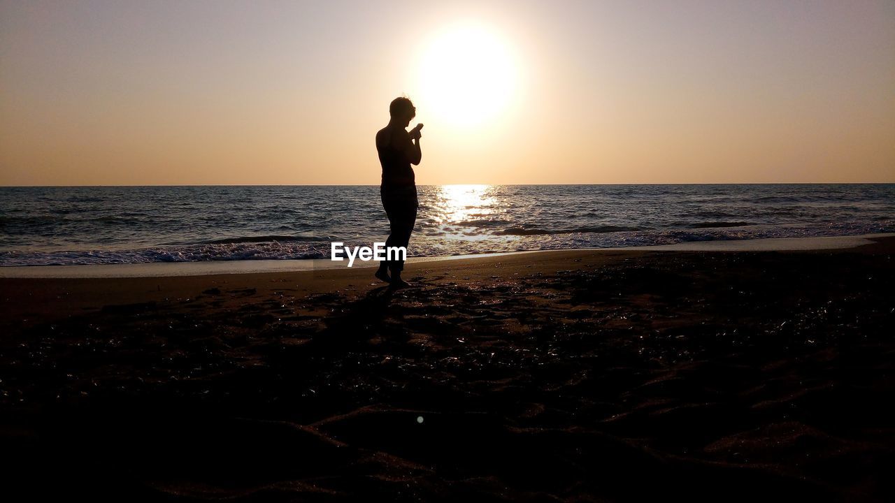 Silhouette woman standing at beach against sky during sunset