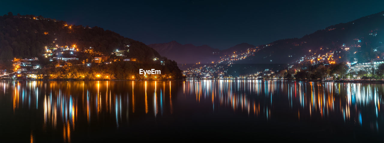 Illuminated city by lake against sky at night