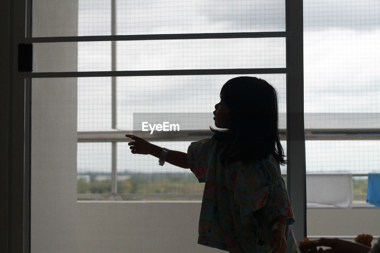 Side view of girl pointing while standing by glass window