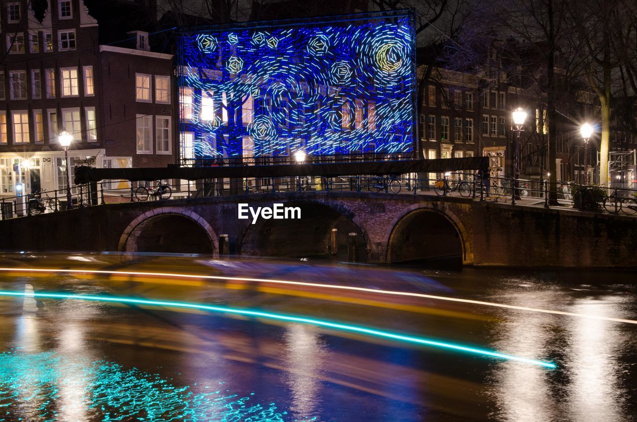 ILLUMINATED BRIDGE OVER RIVER BY CITY BUILDINGS