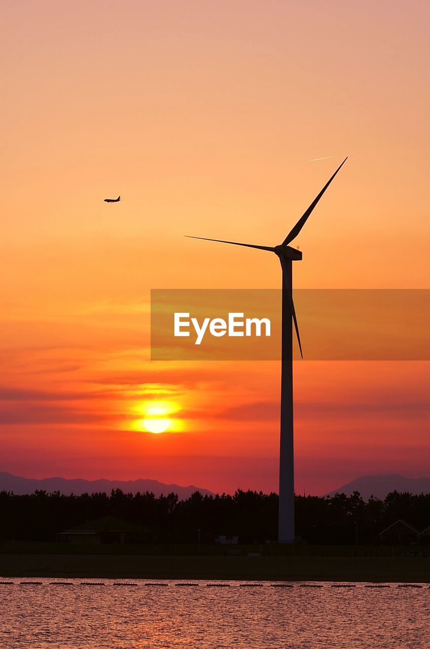 Windmill by lake against sky during sunset