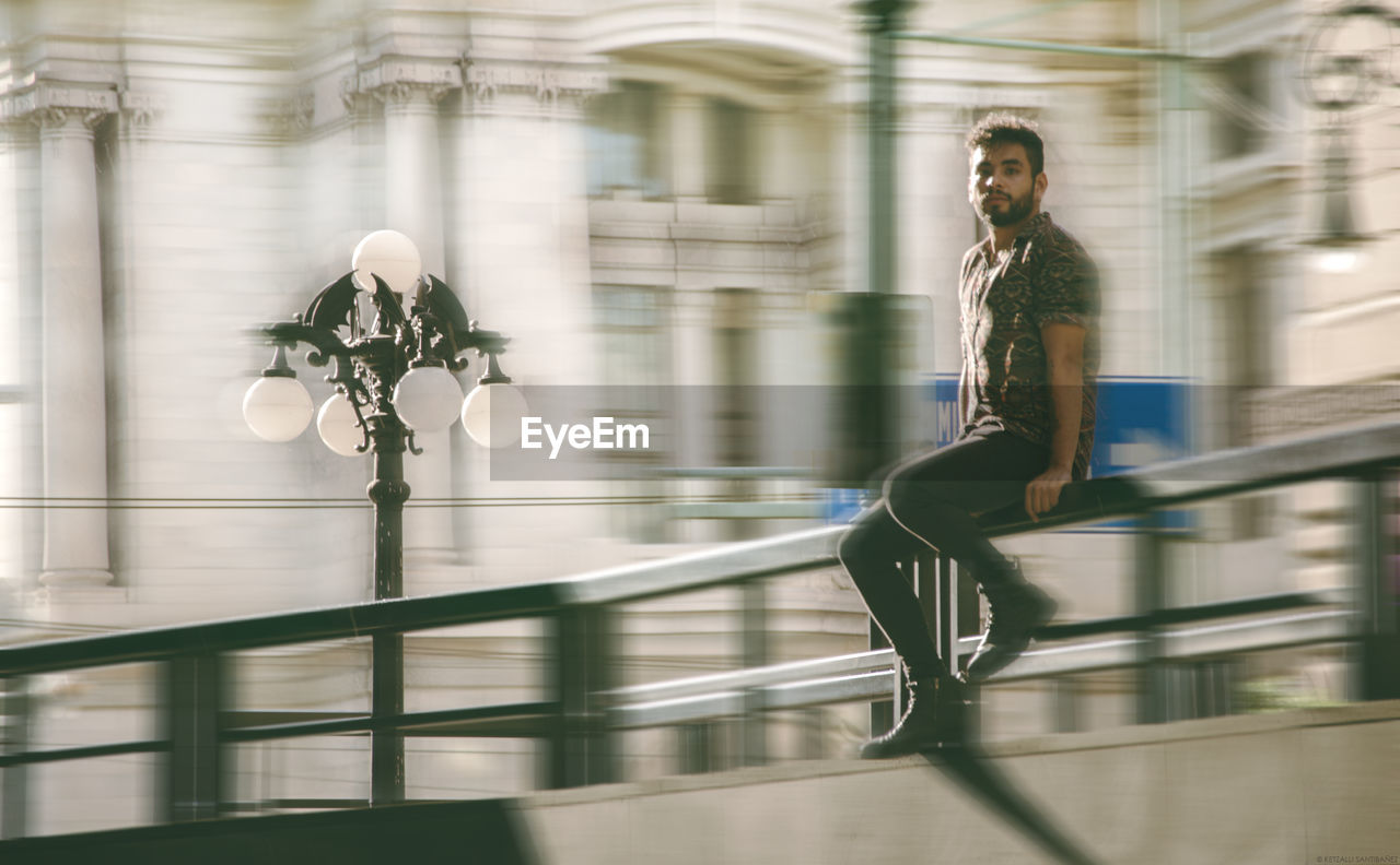 Blurred motion of man sitting on railing by road in city