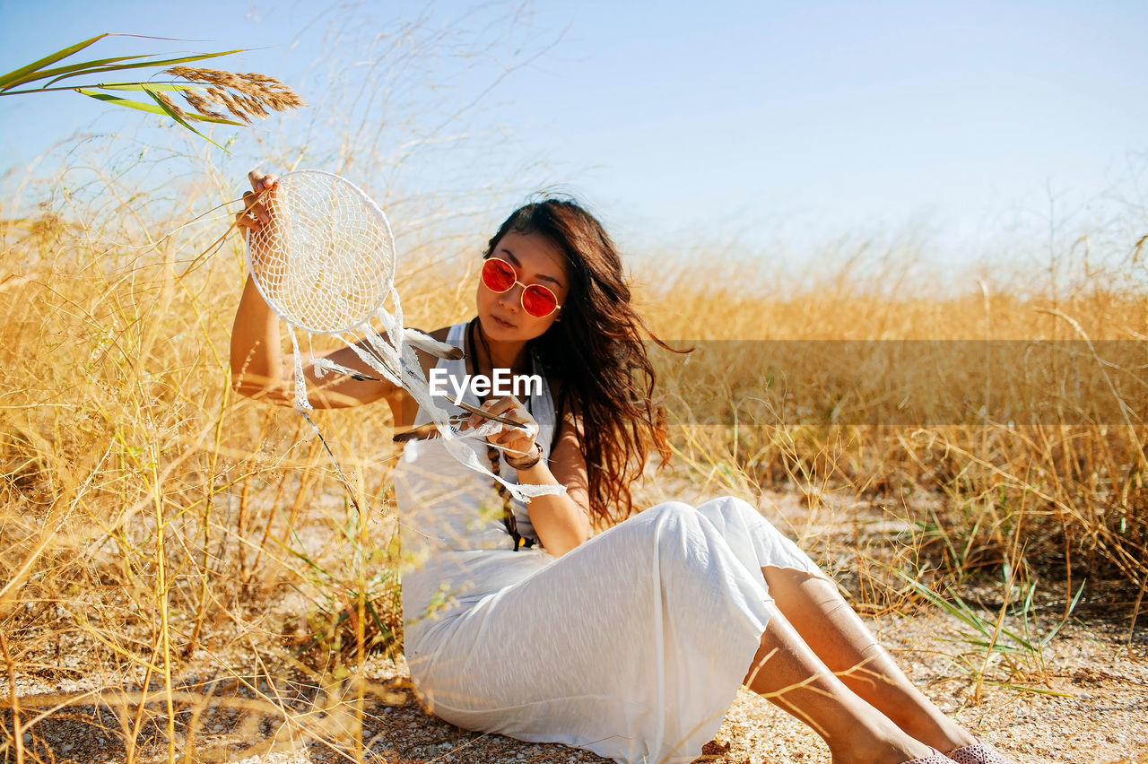 Asian boho chic in long white dress holding dream catcher in field with dry grass.