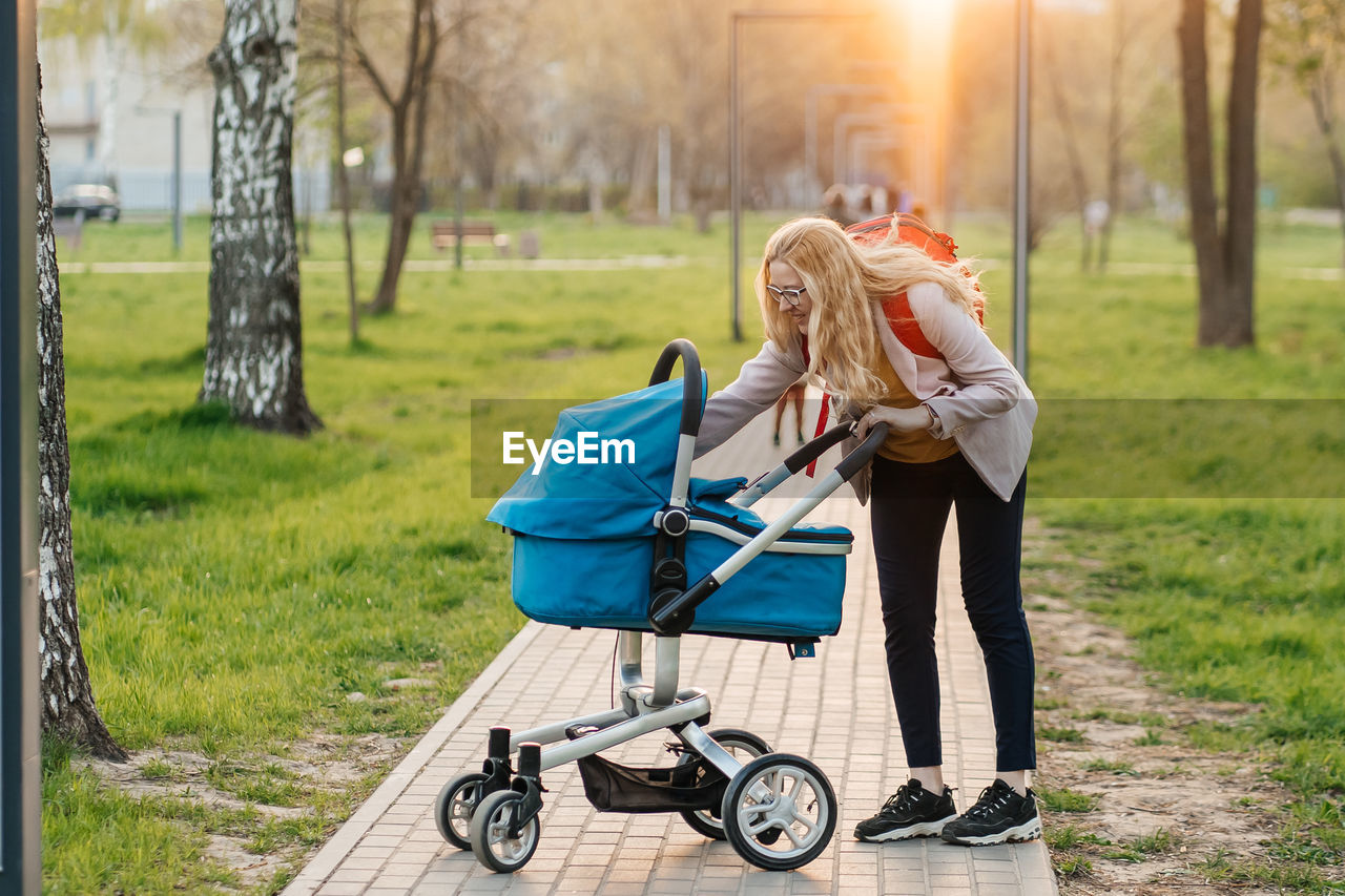 women, baby carriage, adult, two people, female, full length, emotion, nature, lifestyles, togetherness, blond hair, grass, sunlight, casual clothing, transportation, plant, happiness, leisure activity, young adult, tree, clothing, park, person, child, outdoors, park - man made space, day, positive emotion, smiling, vehicle, pushing, childhood, footpath, love, relaxation, bonding, friendship, spring