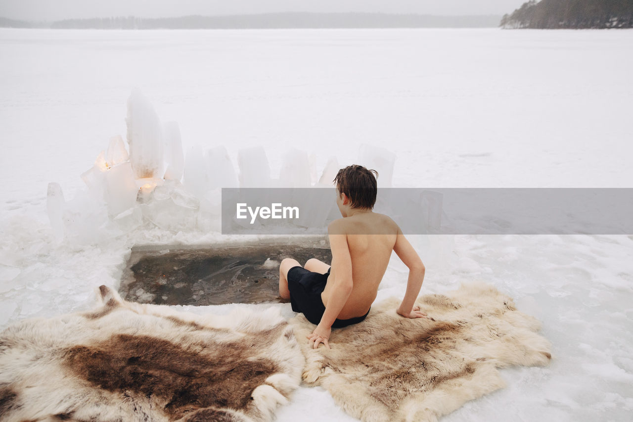 Rear view of shirtless boy sitting with legs in cold water at frozen lake