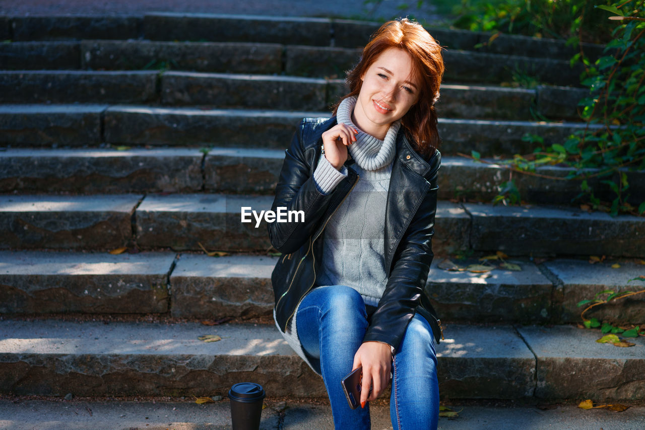  red-haired girl, dressed in a leather jacket with jeans, sits on the steps with a glass of coffee.