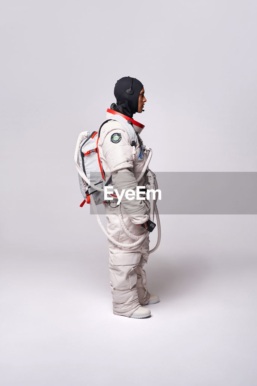 Full body side view of colombian male astronaut in spacesuit with helmet in hand standing in studio against white background and looking away