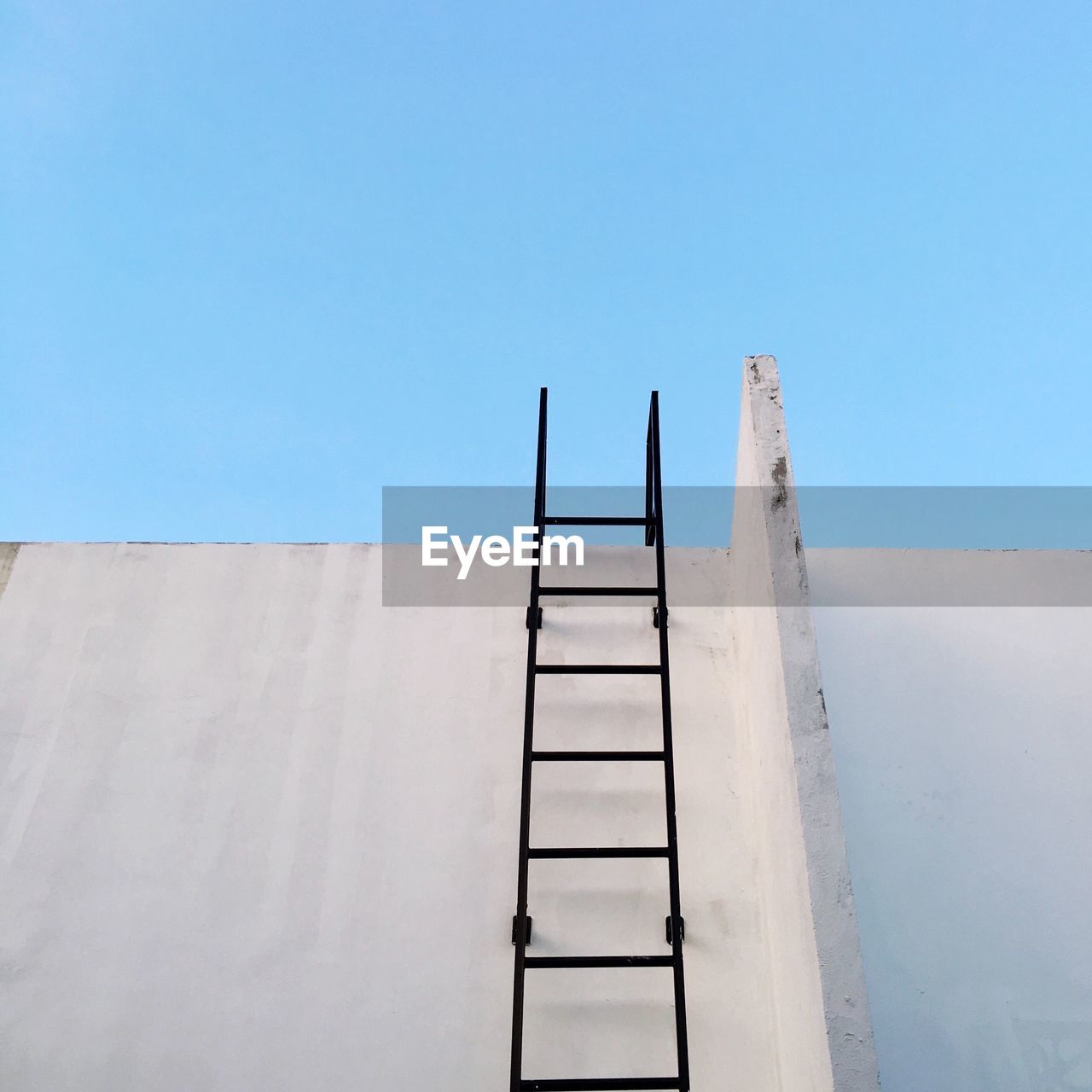 Low angle view of ladder on concrete wall against clear blue sky