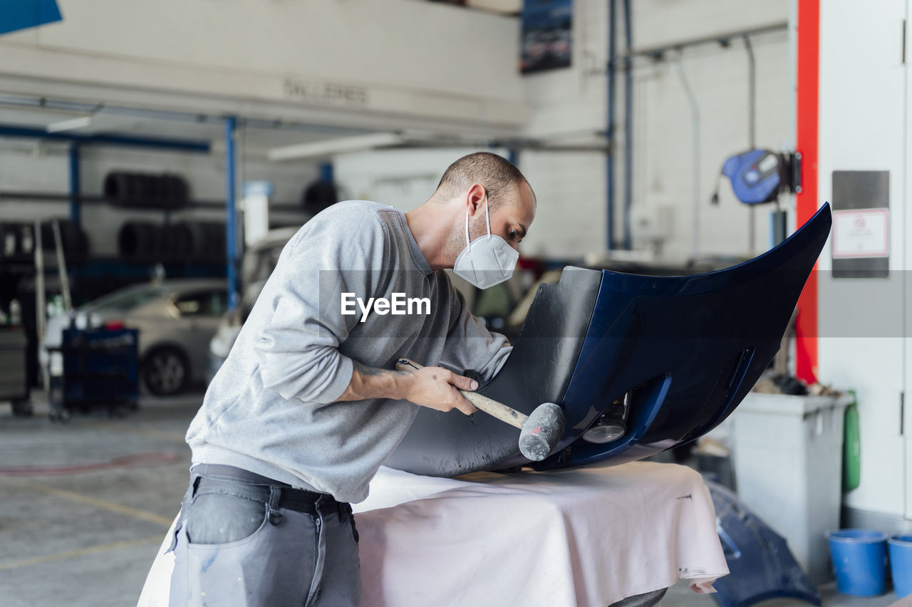 Repairman wearing protective face mask hammering on car body in workshop