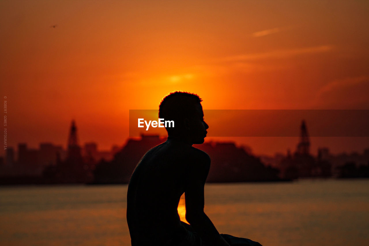 Silhouette man sitting at beach against orange sky